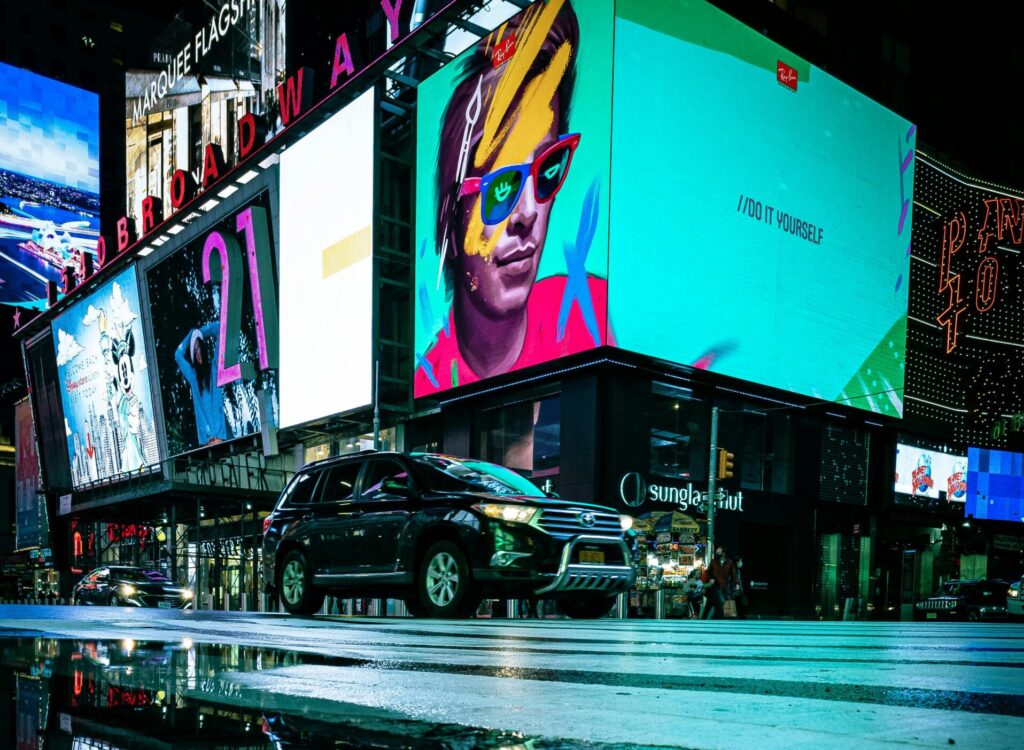 A dynamic scene of illuminated billboards reflected in a puddle on a bustling city street at night.